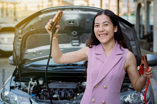 Young beautiful Asian woman holding jumper cable for recharge the battery car. Beautiful woman in purple dressed smiling showing battery cable copper wire while standing in front of by her bronze car.