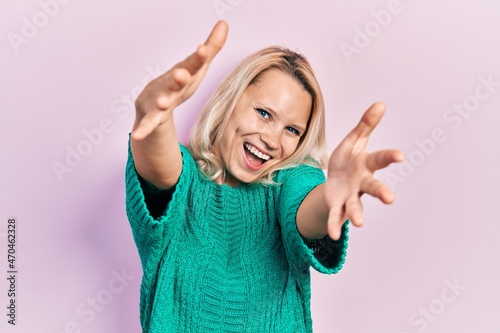 Beautiful caucasian blonde woman wearing casual winter sweater looking at the camera smiling with open arms for hug. cheerful expression embracing happiness.