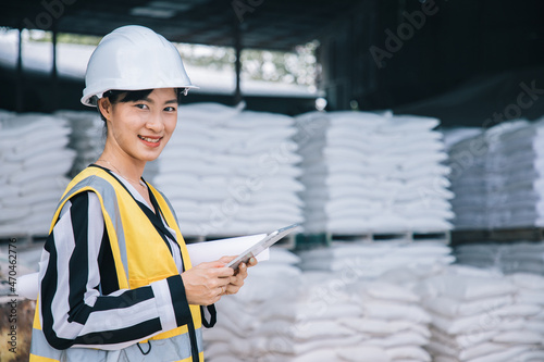 Portrait of beautiful woman engineering using tablet with wear hardhat in front of calcium hydroxide factory. Back view of products in sack of modern safety factory.