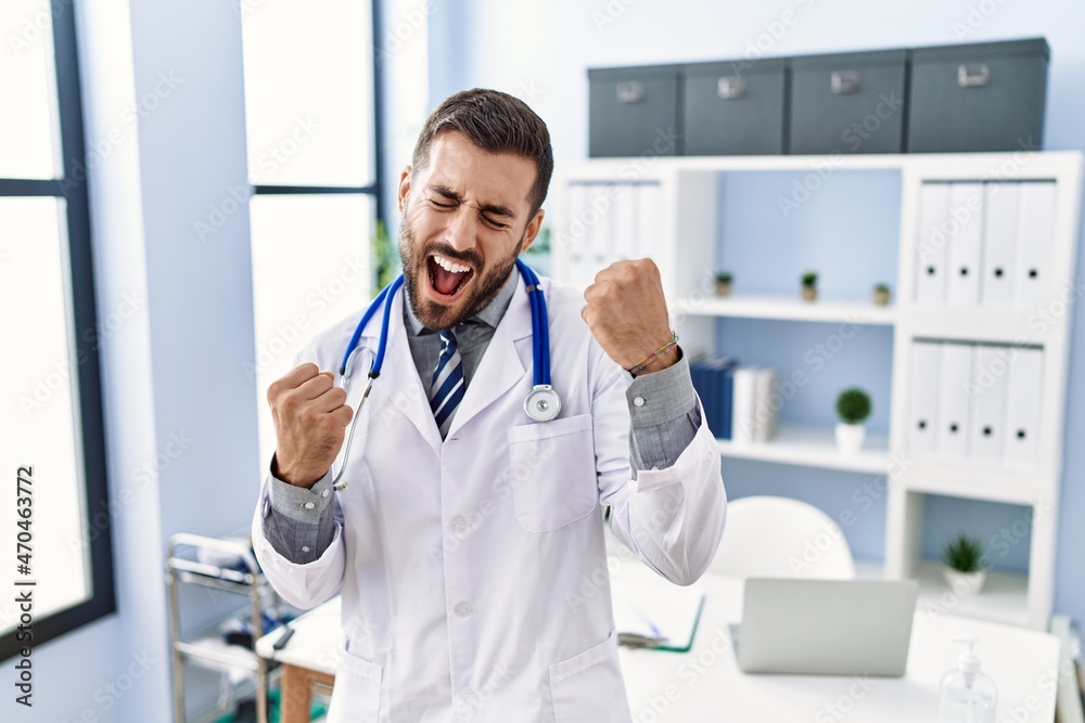 Handsome hispanic man wearing doctor uniform and stethoscope at medical clinic very happy and excited doing winner gesture with arms raised, smiling and screaming for success. celebration concept.