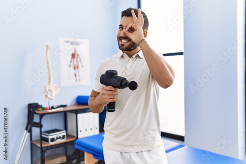 Handsome hispanic man holding therapy massage gun at physiotherapy center smiling happy doing ok sign with hand on eye looking through fingers