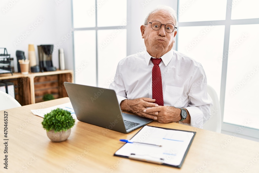 Senior man working at the office using computer laptop puffing cheeks with funny face. mouth inflated with air, crazy expression. Senior man working at the office using computer laptop puffing cheeks with funny face. mouth inflated with air, crazy expression.