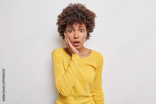 Frightened shocked woman trembles from fear stares with speechless expression keeps hand on cheek cannot believe in something amazing wears casual yellow jumper isolated over white background