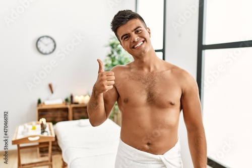 Young hispanic man standing shirtless at spa center doing happy thumbs up gesture with hand. approving expression looking at the camera showing success.
