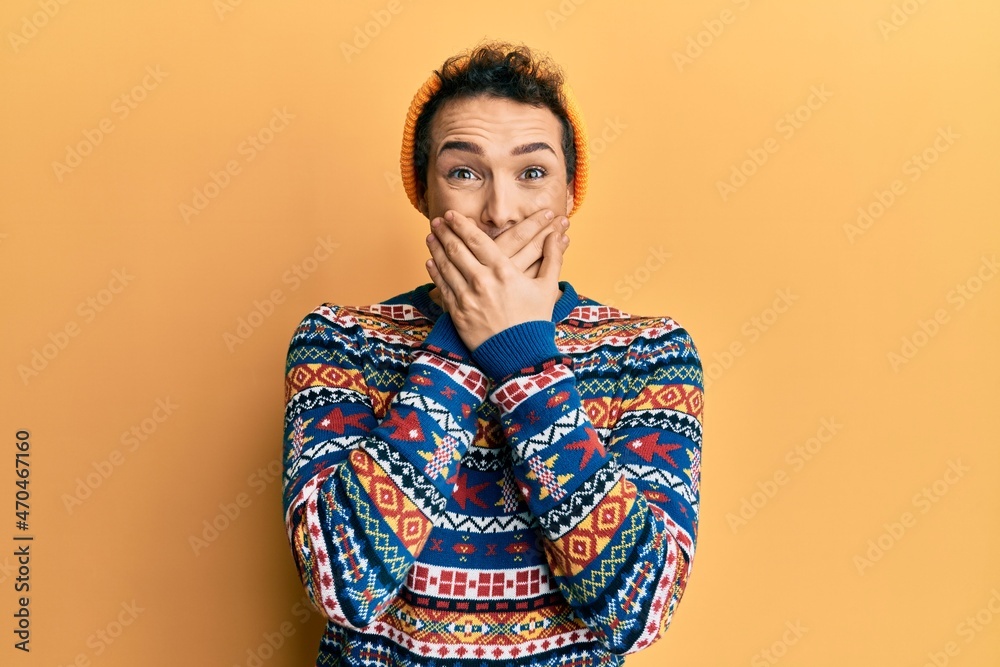 Young handsome man wearing wool hat and colorful sweater shocked covering mouth with hands for mistake. secret concept. Young handsome man wearing wool hat and colorful sweater shocked covering mouth with hands for mistake. secret concept.