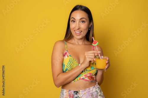 Young smiling woman in summer clothing drinking juice isolated over yellow background.