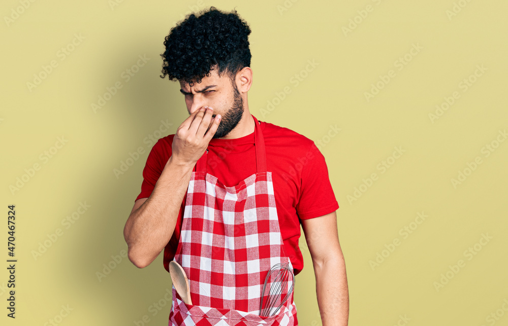 Young arab man with beard wearing cook apron smelling something stinky and disgusting, intolerable smell, holding breath with fingers on nose. bad smell Young arab man with beard wearing cook apron smelling something stinky and disgusting, intolerable smell, holding breath with fingers on nose. bad smell