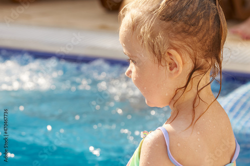 Little cute girl in rainbow swimsuit sitting on pool and smiling in sun bright