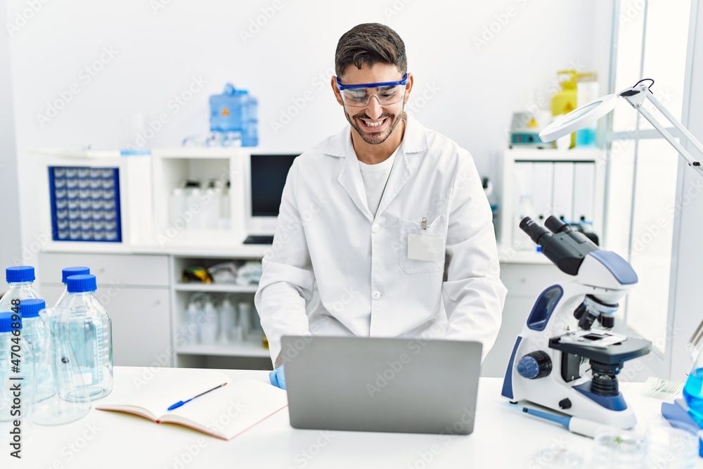 Young hispanic man working at scientist laboratory with laptop looking positive and happy standing and smiling with a confident smile showing teeth Young hispanic man working at scientist laboratory with laptop looking positive and happy standing and smiling with a confident smile showing teeth