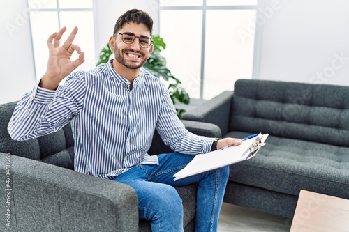 Fototapeta Naklejka Na Ścianę i Meble -  Young psychologist man at consultation office smiling positive doing ok sign with hand and fingers. successful expression.