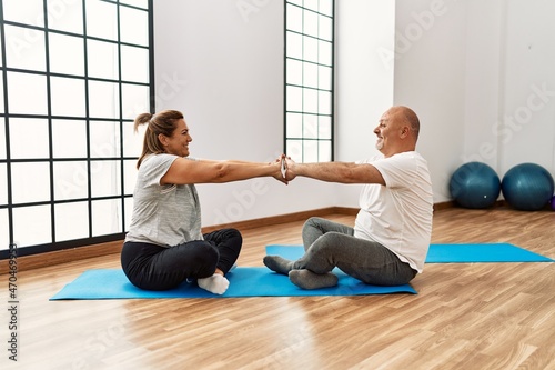 Middle age hispanic couple sitting on yoga mat holding hands at sport center.