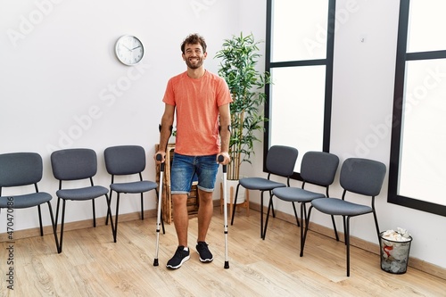Young hispanic man walking using crutches at clinic waiting room.
