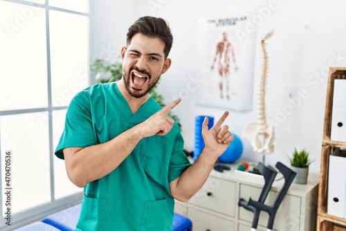 Young man with beard working at pain recovery clinic smiling and looking at the camera pointing with two hands and fingers to the side.