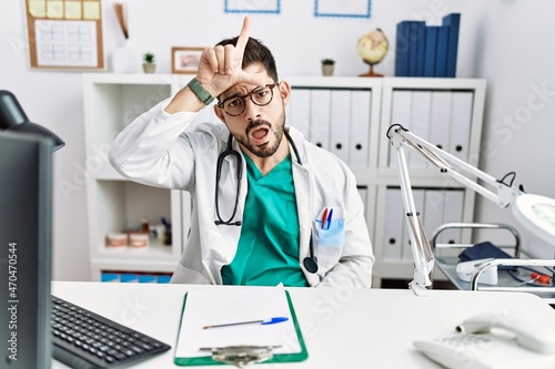 Young man with beard wearing doctor uniform and stethoscope at the clinic making fun of people with fingers on forehead doing loser gesture mocking and insulting.