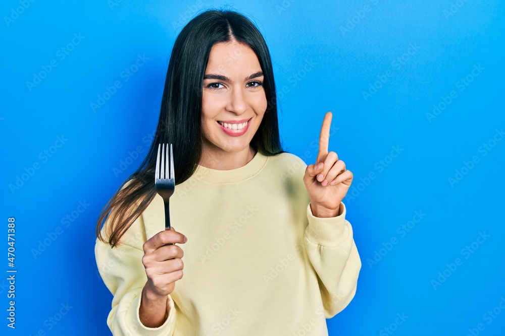 Young brunette woman holding one silver fork smiling with an idea or question pointing finger with happy face, number one Young brunette woman holding one silver fork smiling with an idea or question pointing finger with happy face, number one