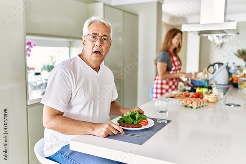 Middle age hispanic couple eating healthy meal at home scared and amazed with open mouth for surprise, disbelief face