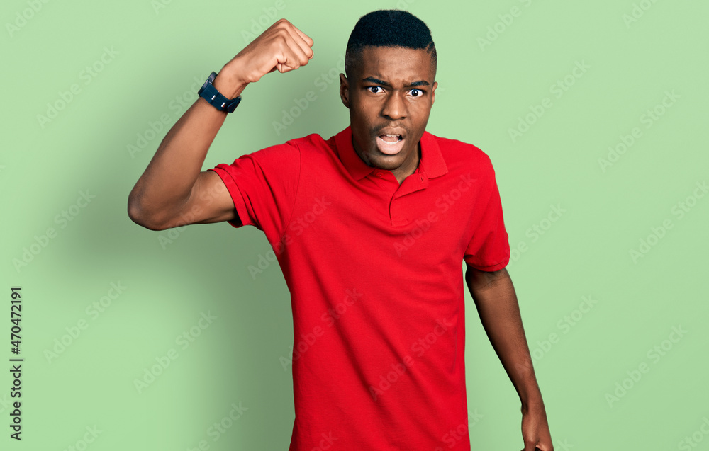 Young african american man wearing casual red t shirt angry and mad raising fist frustrated and furious while shouting with anger. rage and aggressive concept.
