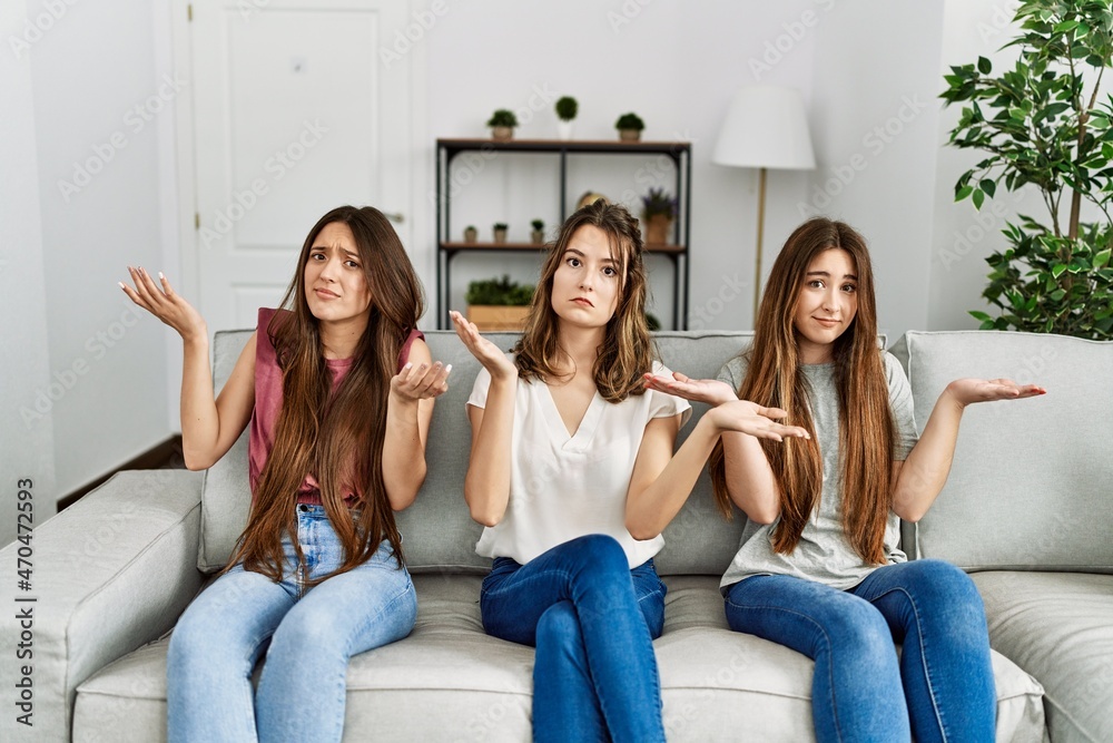 Group of three hispanic girls sitting on the sofa at home clueless and confused expression with arms and hands raised. doubt concept. Group of three hispanic girls sitting on the sofa at home clueless and confused expression with arms and hands raised. doubt concept.