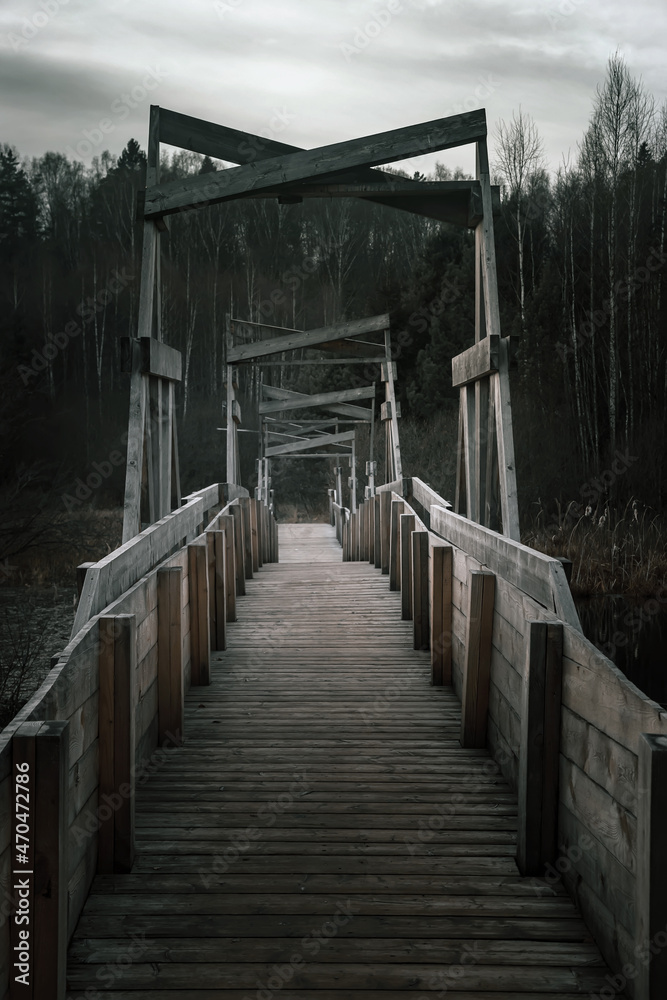 Obraz premium Old and gloomy wooden bridge over the river. Detachment in the water. Gloomy and mysterious forest. Cloudy sky. Wooden structure of an old abandoned bridge.