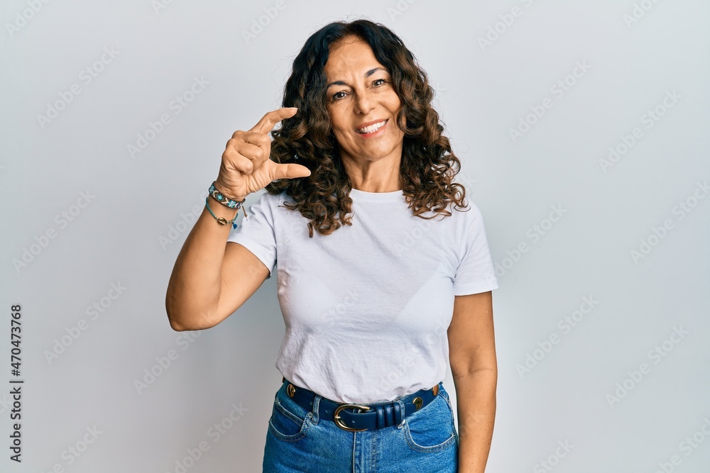 Middle age hispanic woman wearing casual white t shirt smiling and confident gesturing with hand doing small size sign with fingers looking and the camera. measure concept. Middle age hispanic woman wearing casual white t shirt smiling and confident gesturing with hand doing small size sign with fingers looking and the camera. measure concept.