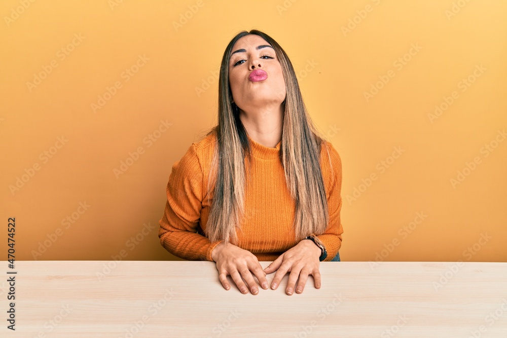 Young hispanic woman wearing casual clothes sitting on the table looking at the camera blowing a kiss on air being lovely and sexy. love expression. Young hispanic woman wearing casual clothes sitting on the table looking at the camera blowing a kiss on air being lovely and sexy. love expression.