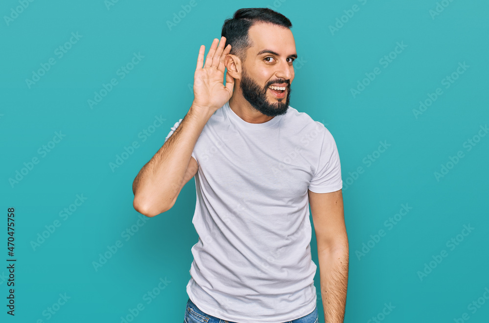 Young man with beard wearing casual white t shirt smiling with hand over ear listening an hearing to rumor or gossip. deafness concept. Young man with beard wearing casual white t shirt smiling with hand over ear listening an hearing to rumor or gossip. deafness concept.