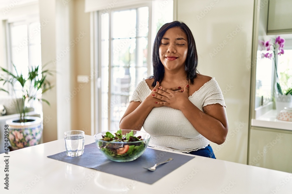 Young hispanic woman eating healthy salad at home smiling with hands on chest with closed eyes and grateful gesture on face. health concept. Young hispanic woman eating healthy salad at home smiling with hands on chest with closed eyes and grateful gesture on face. health concept.