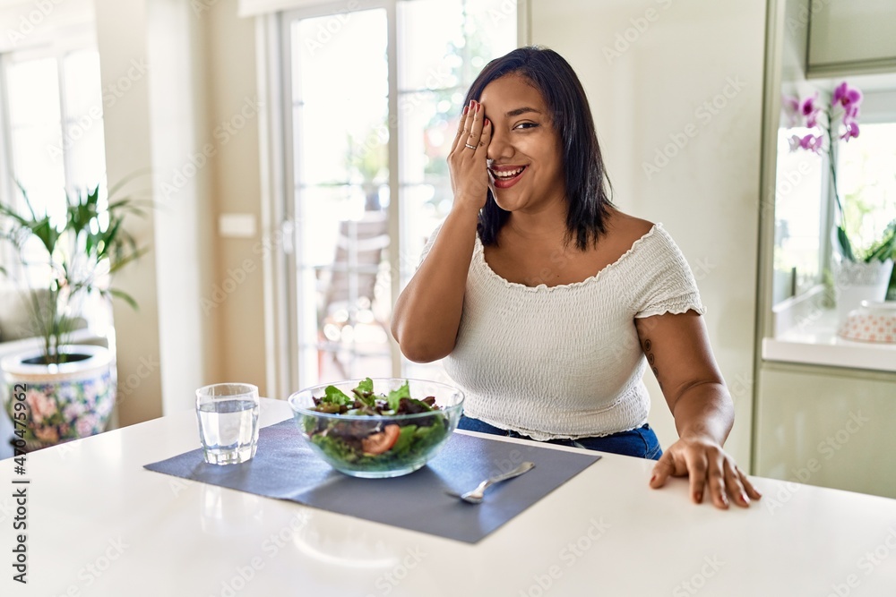 Young hispanic woman eating healthy salad at home covering one eye with hand, confident smile on face and surprise emotion.