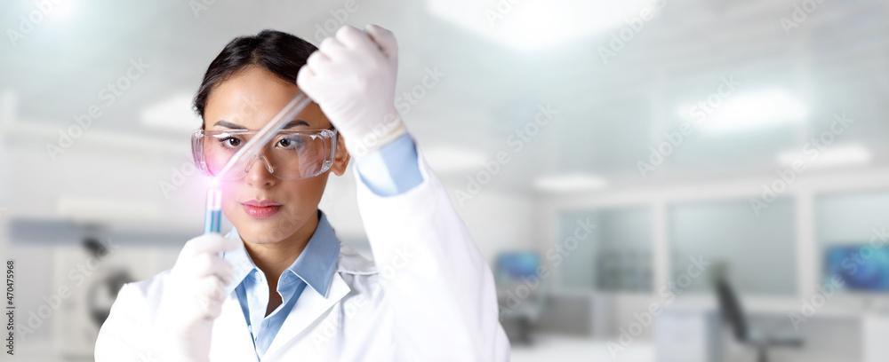 Naklejka premium Young focused female chemist in lab coat, gloves and protective eyeglass dropping a blue substance into a test tube with a pipette in laboratory. Chemical, pharmaceutical or medical research concept.