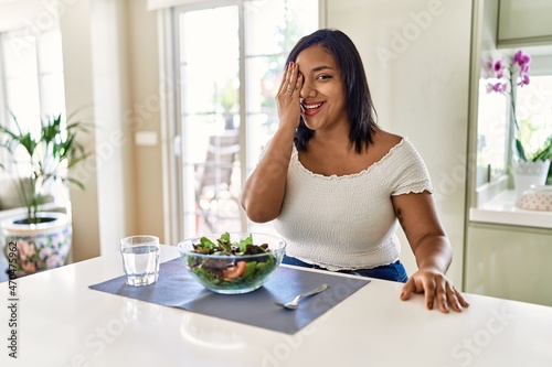 Young hispanic woman eating healthy salad at home covering one eye with hand  confident smile on face and surprise emotion.