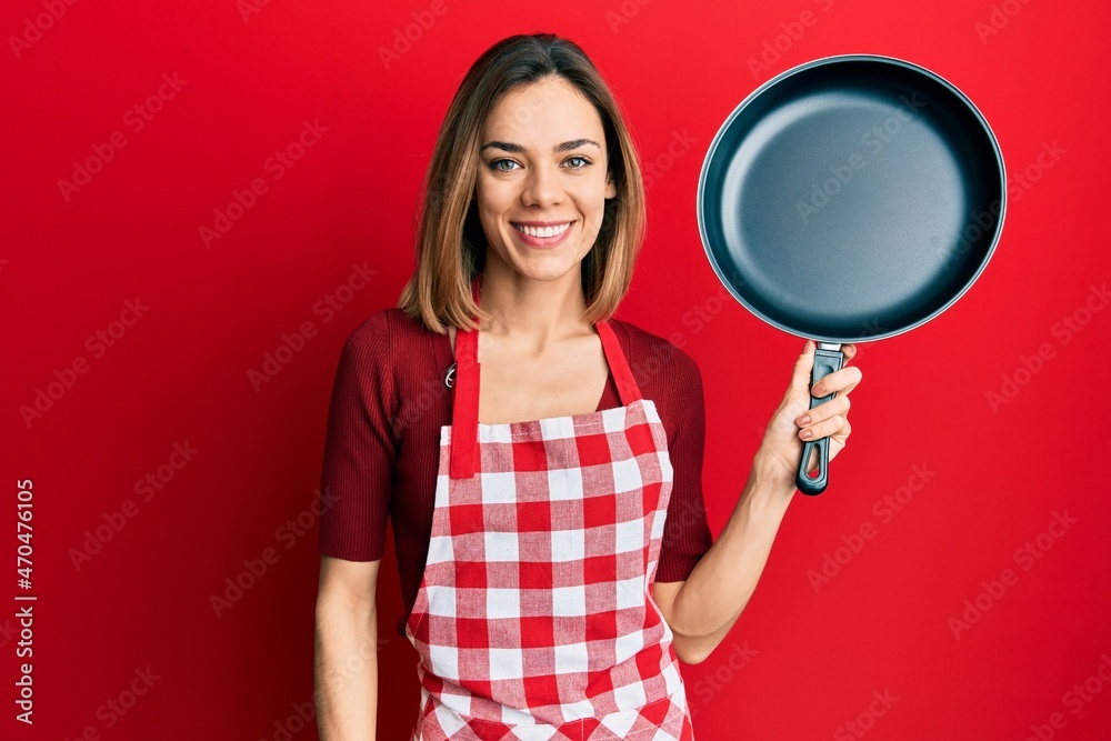 Young caucasian blonde woman wearing cook apron holding pan looking positive and happy standing and smiling with a confident smile showing teeth
