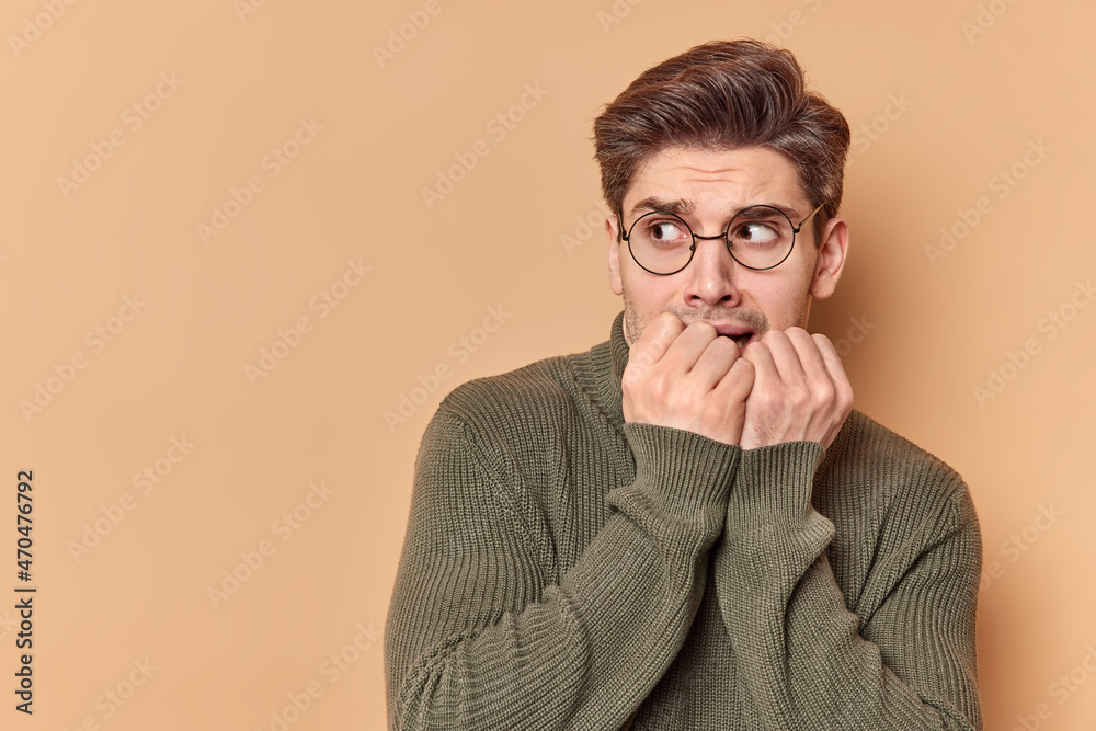 Scared anxious young man bites finger nails looks nervously away being afraid of something wears round spectacles casual jumper has anxiety isolated over beige studio wall with copy space area Scared anxious young man bites finger nails looks nervously away being afraid of something wears round spectacles casual jumper has anxiety isolated over beige studio wall with copy space area