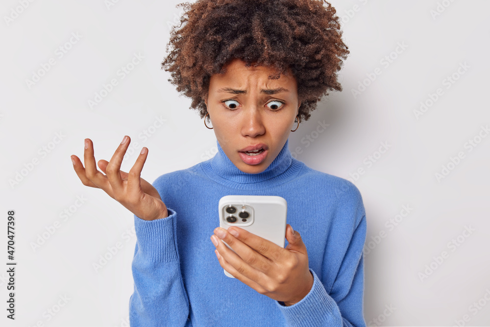 Indignant woman stares at smartphone screen raises palm reacts on embarrassed news has puzzled expression wears blue turtleneck isolated over white background. Shocked female telephone user. Indignant woman stares at smartphone screen raises palm reacts on embarrassed news has puzzled expression wears blue turtleneck isolated over white background. Shocked female telephone user.