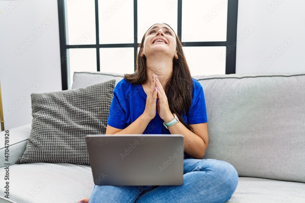 Fototapeta premium Young brunette woman using laptop at home begging and praying with hands together with hope expression on face very emotional and worried. begging.