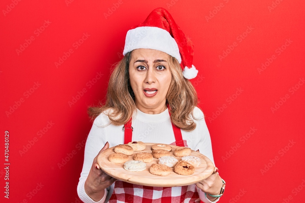 Middle age caucasian woman wearing baker apron and christmas hat holding tray with biscuits clueless and confused expression. doubt concept. Middle age caucasian woman wearing baker apron and christmas hat holding tray with biscuits clueless and confused expression. doubt concept.