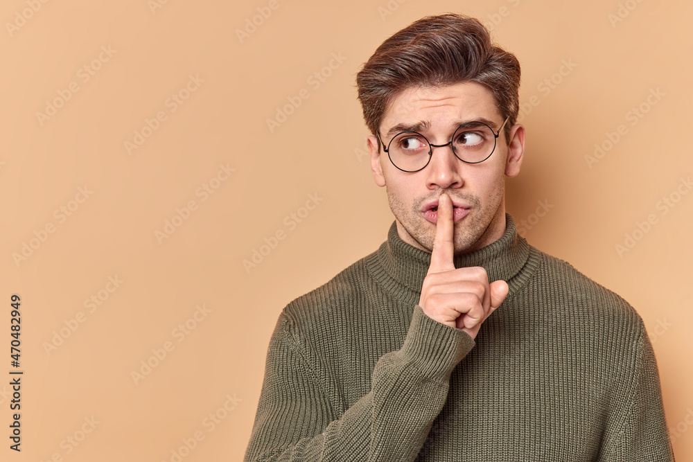 Be quiet. Serious mysterious man presses index finger to lips shows taboo sign tells to keep silence shushing indoor wears casual clothes looks away isolated over beige background blank copy space - obrazy, fototapety, plakaty 
