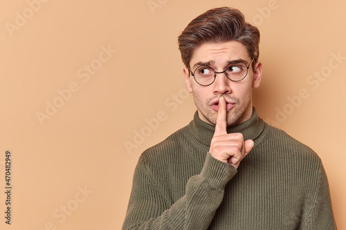 Be quiet. Serious mysterious man presses index finger to lips shows taboo sign tells to keep silence shushing indoor wears casual clothes looks away isolated over beige background blank copy space