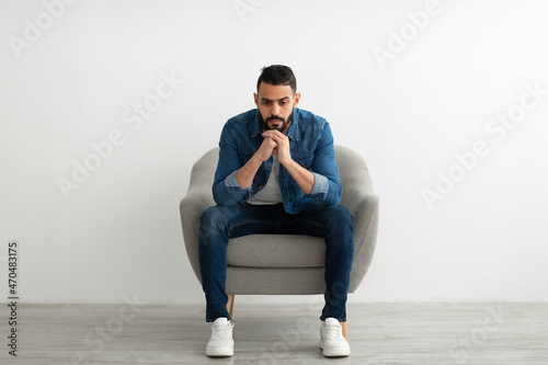 Arab man sitting in armchair with dull face expression, feeling sad or tired, having problem, suffering from depression