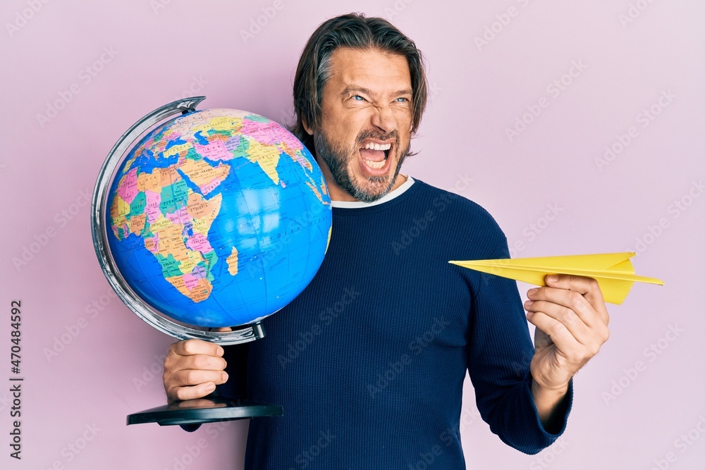 Fototapeta premium Middle age handsome man holding paper plane and world ball angry and mad screaming frustrated and furious, shouting with anger. rage and aggressive concept.