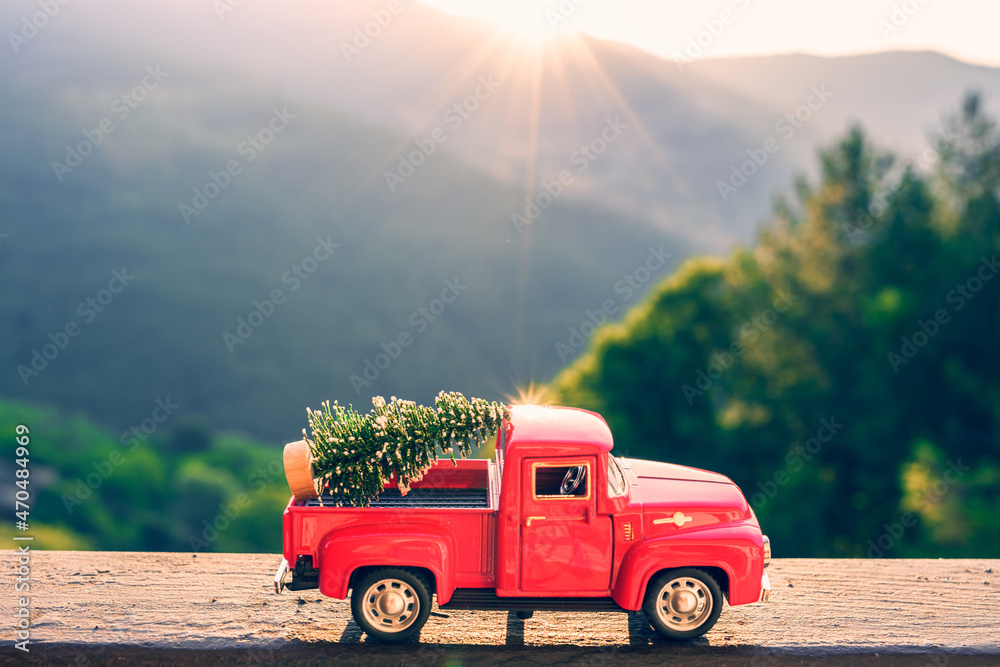 Red retro small car carrying christmas tree in the mountain in the sunbeams of sunset. Winter holiday concept