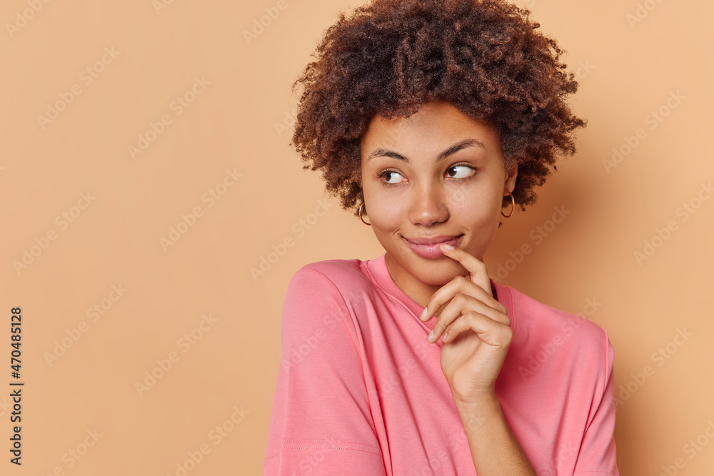 Portrait of good looking curly haired young woman looks left with dreamy expression smiles gently wears casual clothes admires something isolated over beige background with blank copy space. Portrait of good looking curly haired young woman looks left with dreamy expression smiles gently wears casual clothes admires something isolated over beige background with blank copy space.