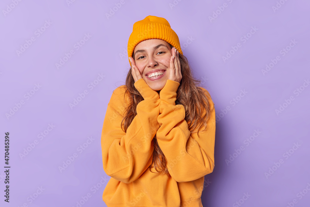 Tender cheerful woman keeps hands on cheeks smiles toothily has glad expression dressed in orange casual jumper and hat isolated over purple background. Pretty millennial girl feels very happy