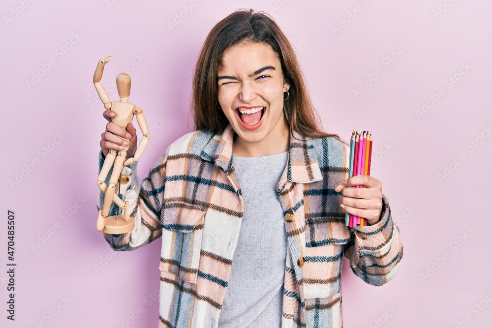 Young caucasian girl holding small wooden manikin and pencils winking looking at the camera with sexy expression, cheerful and happy face. Young caucasian girl holding small wooden manikin and pencils winking looking at the camera with sexy expression, cheerful and happy face.