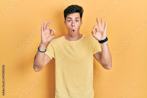 Young hispanic man wearing casual yellow t shirt looking surprised and shocked doing ok approval symbol with fingers. crazy expression