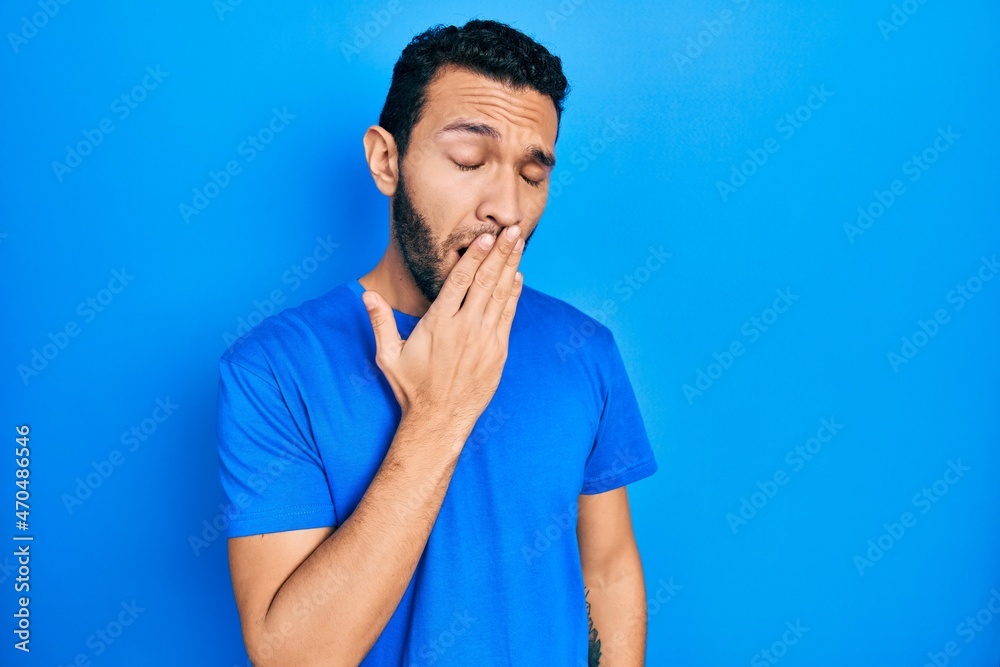 Hispanic man with beard wearing casual blue t shirt bored yawning tired covering mouth with hand. restless and sleepiness. Hispanic man with beard wearing casual blue t shirt bored yawning tired covering mouth with hand. restless and sleepiness.