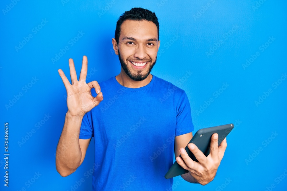 Hispanic man with beard using touchpad device doing ok sign with fingers, smiling friendly gesturing excellent symbol Hispanic man with beard using touchpad device doing ok sign with fingers, smiling friendly gesturing excellent symbol