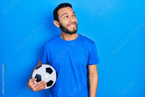 Hispanic man with beard holding soccer ball smiling looking to the side and staring away thinking.