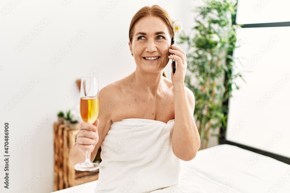 Middle age caucasian woman wearing towel talking on the smartphone sitting on massage board at beauty center Middle age caucasian woman wearing towel talking on the smartphone sitting on massage board at beauty center
