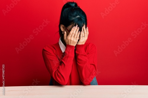 Young brunette woman with bangs wearing glasses sitting on the table with sad expression covering face with hands while crying. depression concept.