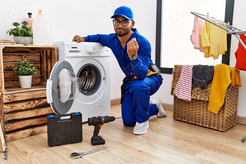 Young indian technician working on washing machine doing money gesture with hands, asking for salary payment, millionaire business